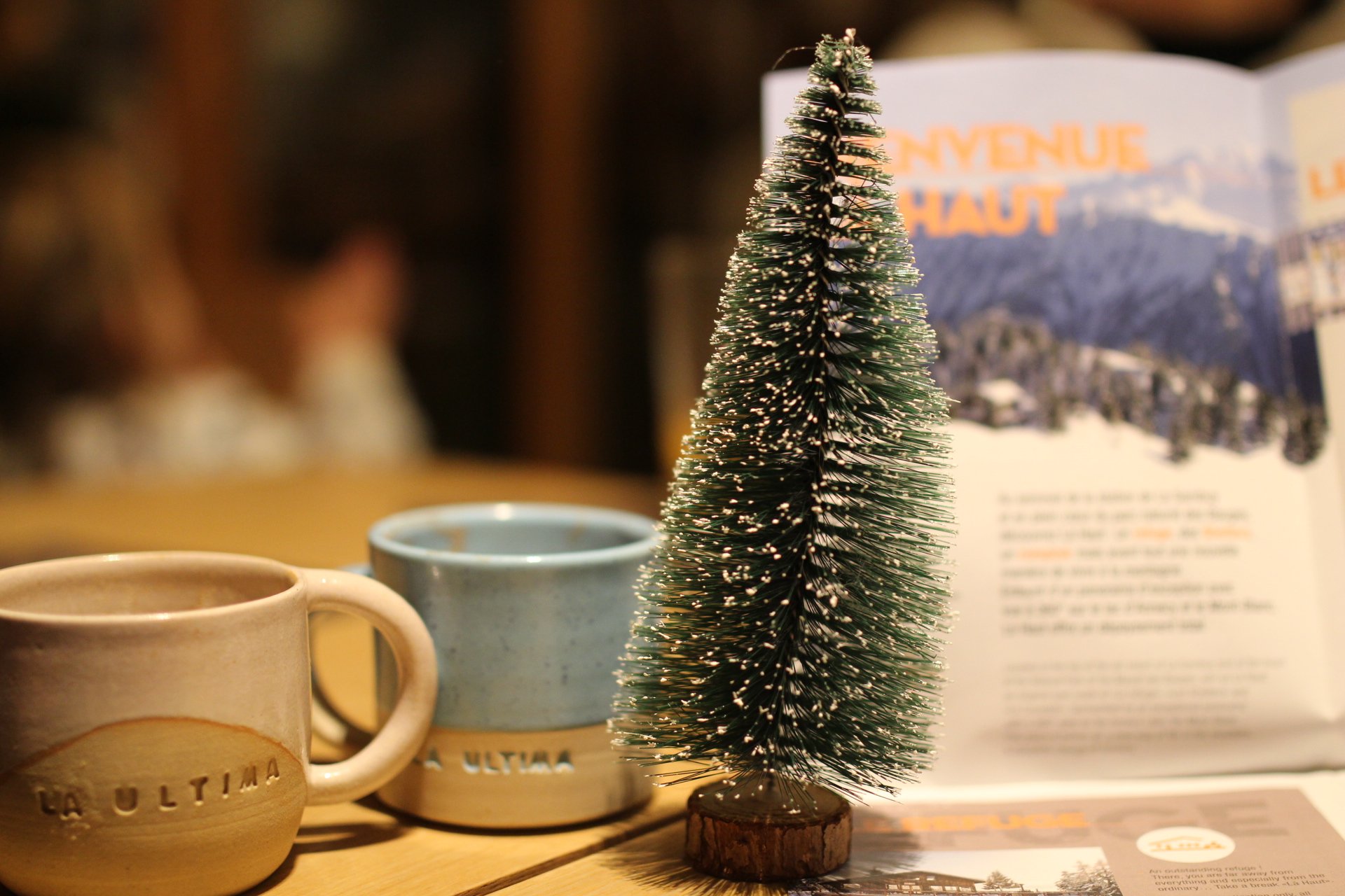 Une table dans un café avec deux tasses, un sapin neige miniature, un carnet de voyage.