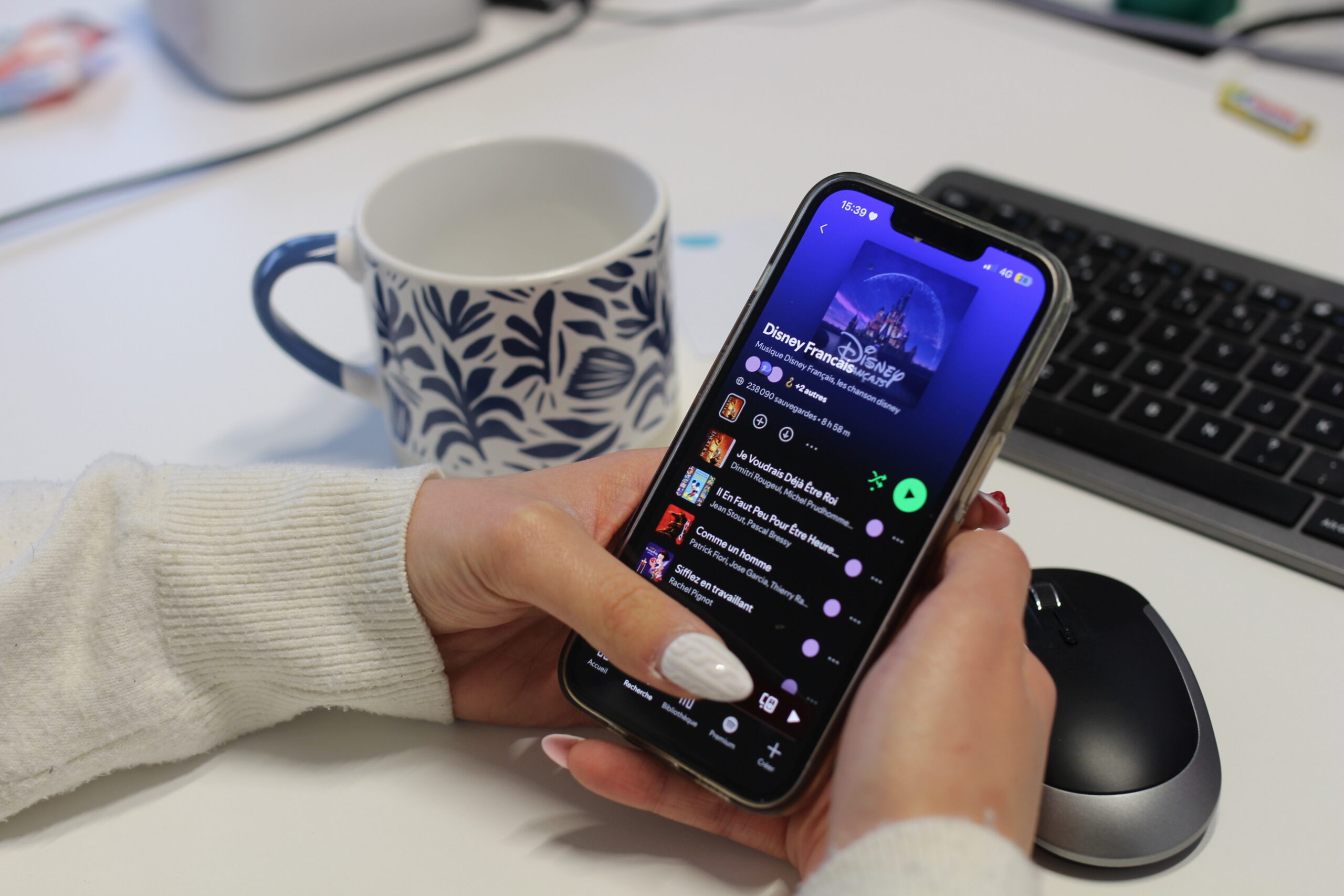 On aperçoit un téléphone type smartphone avec une application et une playlist de blind test dans une salle avec une tasse de café derrière.