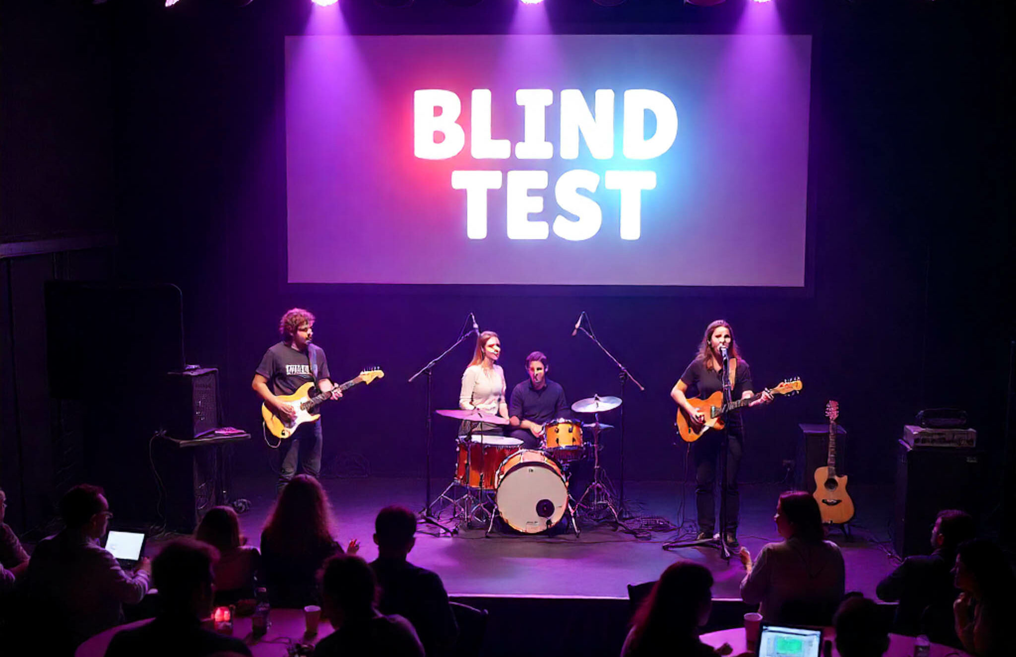 Une scène de concert avec deux guitares, une batterie, une chanteuse. En arrière-plan, projeté sur fond blanc, l'inscription de Blind Test.