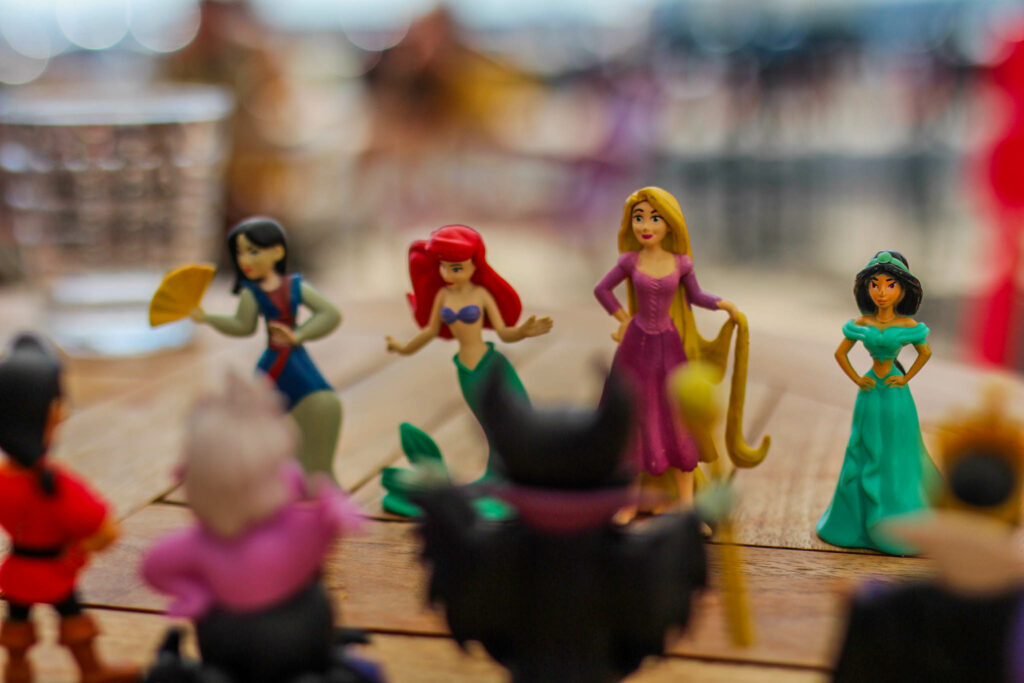 Four Disney princess figurines on a wooden table. From behind, in the foreground, four Disney Villain figurines.