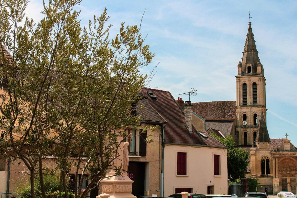 un olivier dans un centre-ville en pierre abritant un monument. Une église en arrière plan et un ciel bleu.