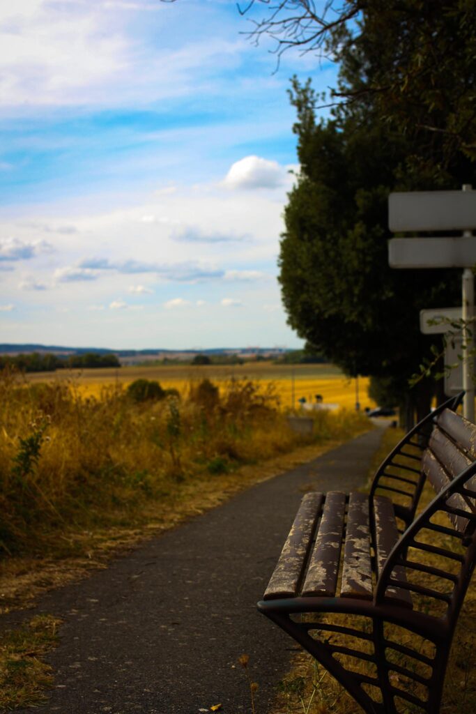 un banc au premier plan avec une voie pour les piétons à côté. Au fond une perspective sur des champs et l'horizon.
