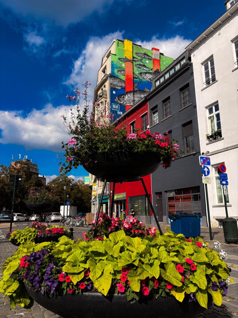 Deux bacs à fleurs superposés et fleuris dans le centre-ville de Bruxelles avec un immeuble street-art en arrière plan.